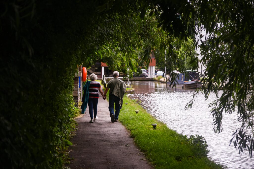 Back view of full length anonymous elderly couple in casual clothes walking along pond and green lawn of park holding hands